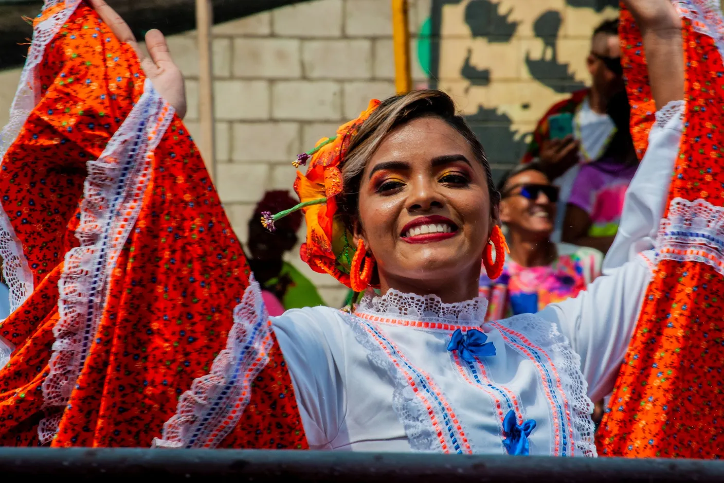 A carnival dancer in a red floral costume and flower headpiece, arms raised mid-dance