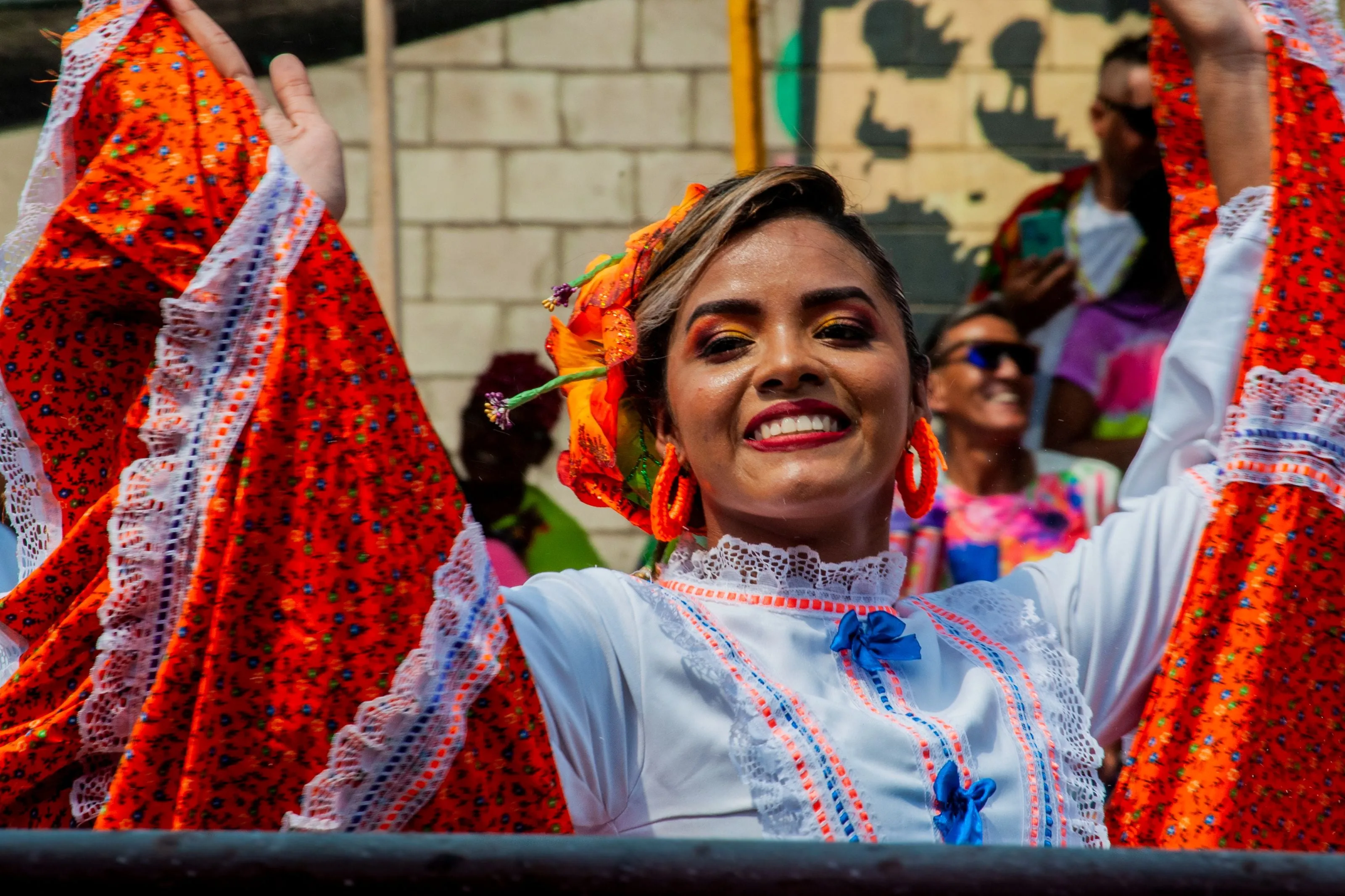 A carnival dancer in a red floral costume and flower headpiece, arms raised mid-dance