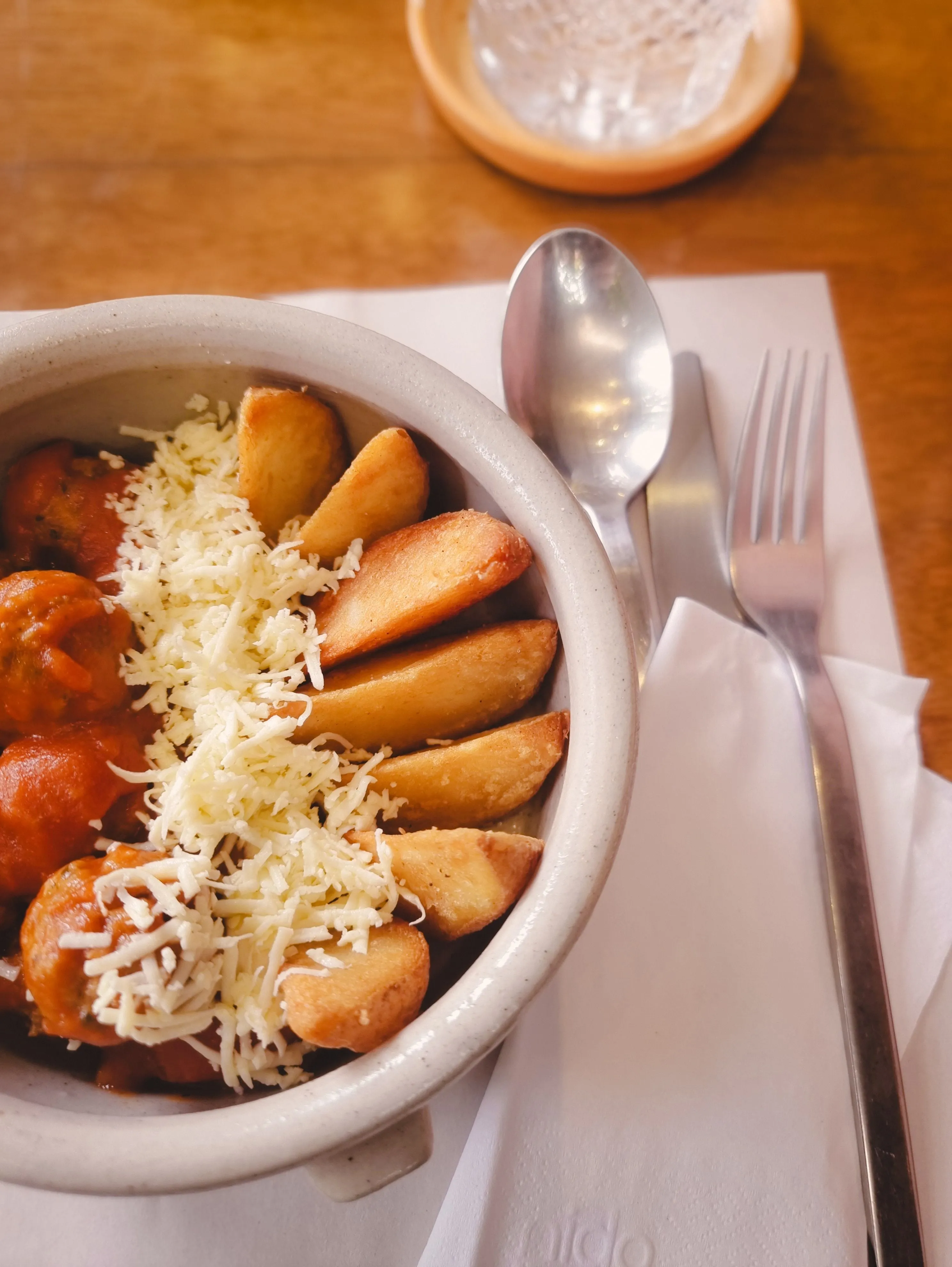 A clay bowl of stew topped with fried potato wedges and grated cheese, with cutlery alongside