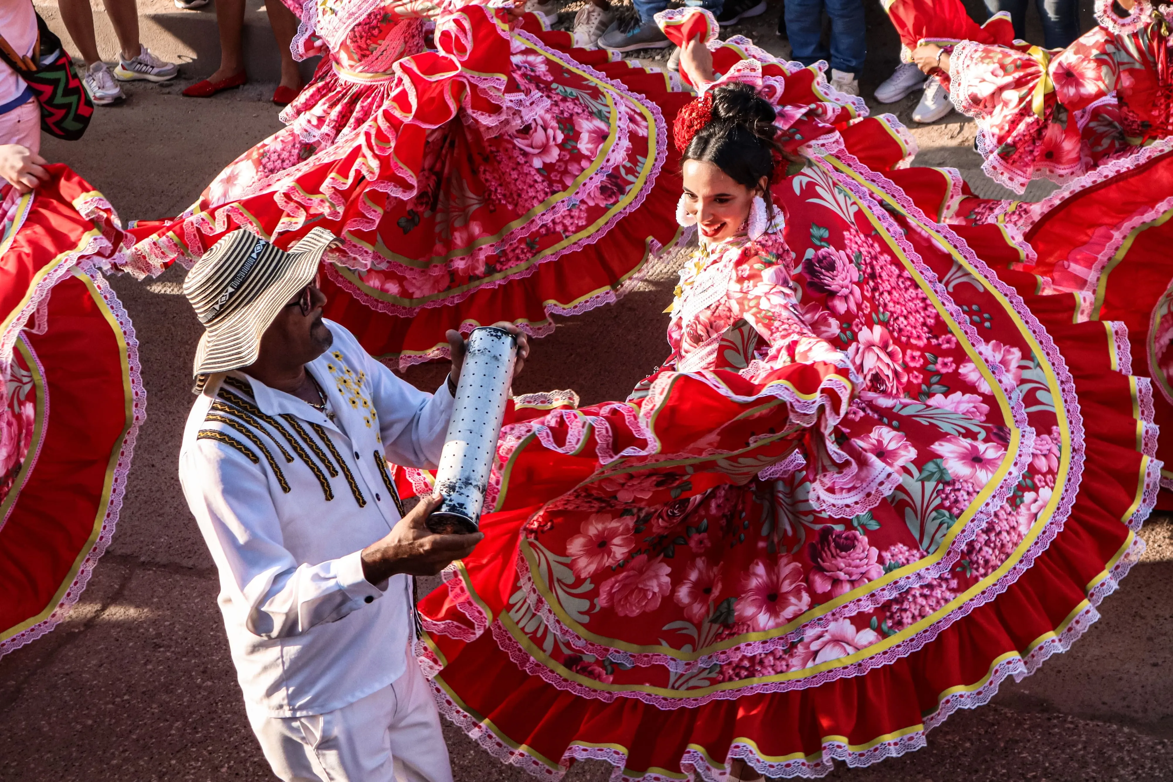 Dancers in wide red and floral skirts mid-turn, with a musician in a sombrero vueltiao playing beside them