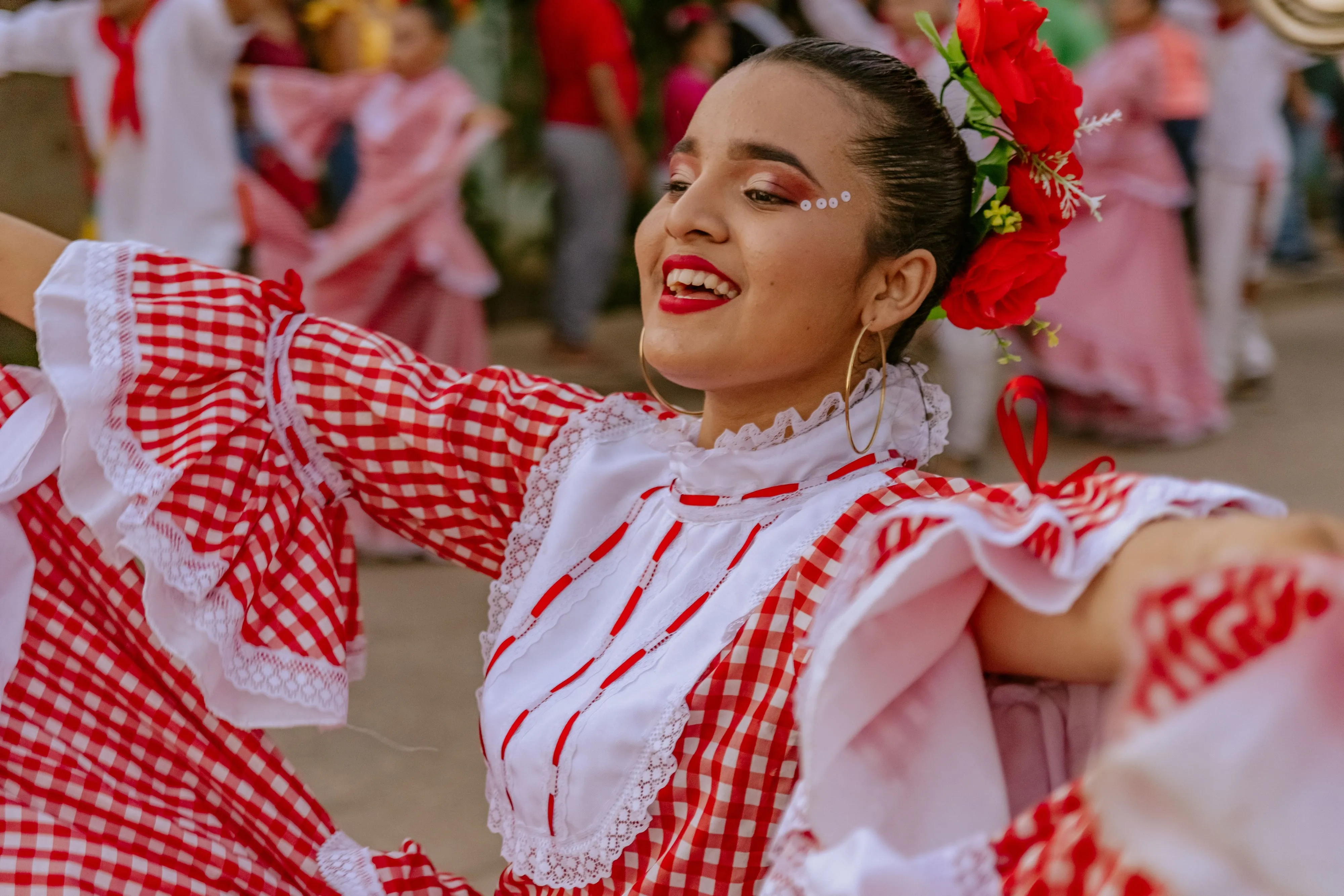 A dancer in a red gingham dress turning mid-step during a street festival, with other dancers behind her