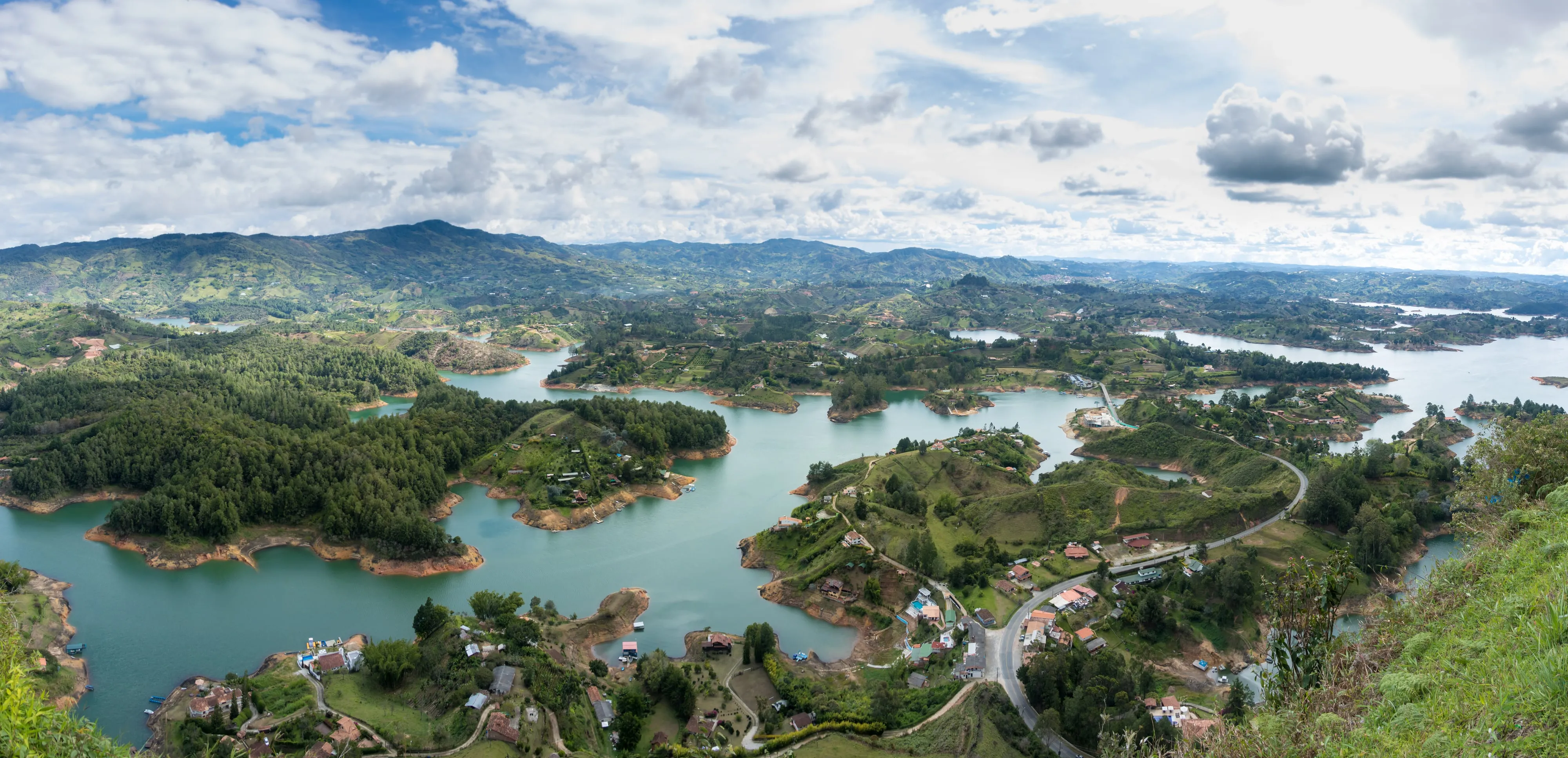 A wide panorama of the Guatapé reservoir, its blue water winding between green islands and hills under a bright sky