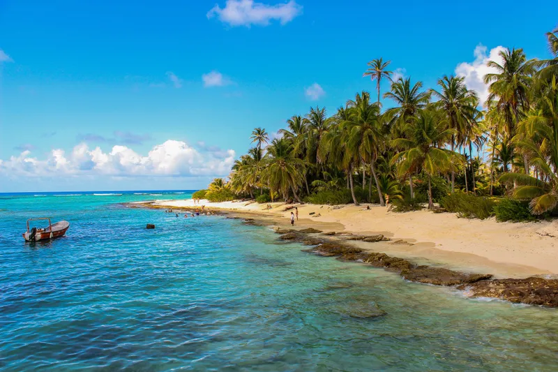 Palms and white sand on a Caribbean cay, the water banding from deep blue through turquoise to pale green over the reef flat